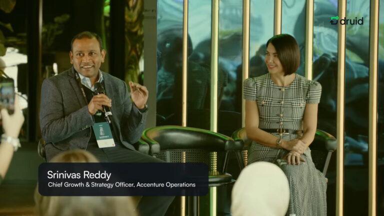 Panel discussion at Symbiosis 4: a speaker holds a microphone and gestures while another panelist listens; audience in the foreground and Druid branding in the background.